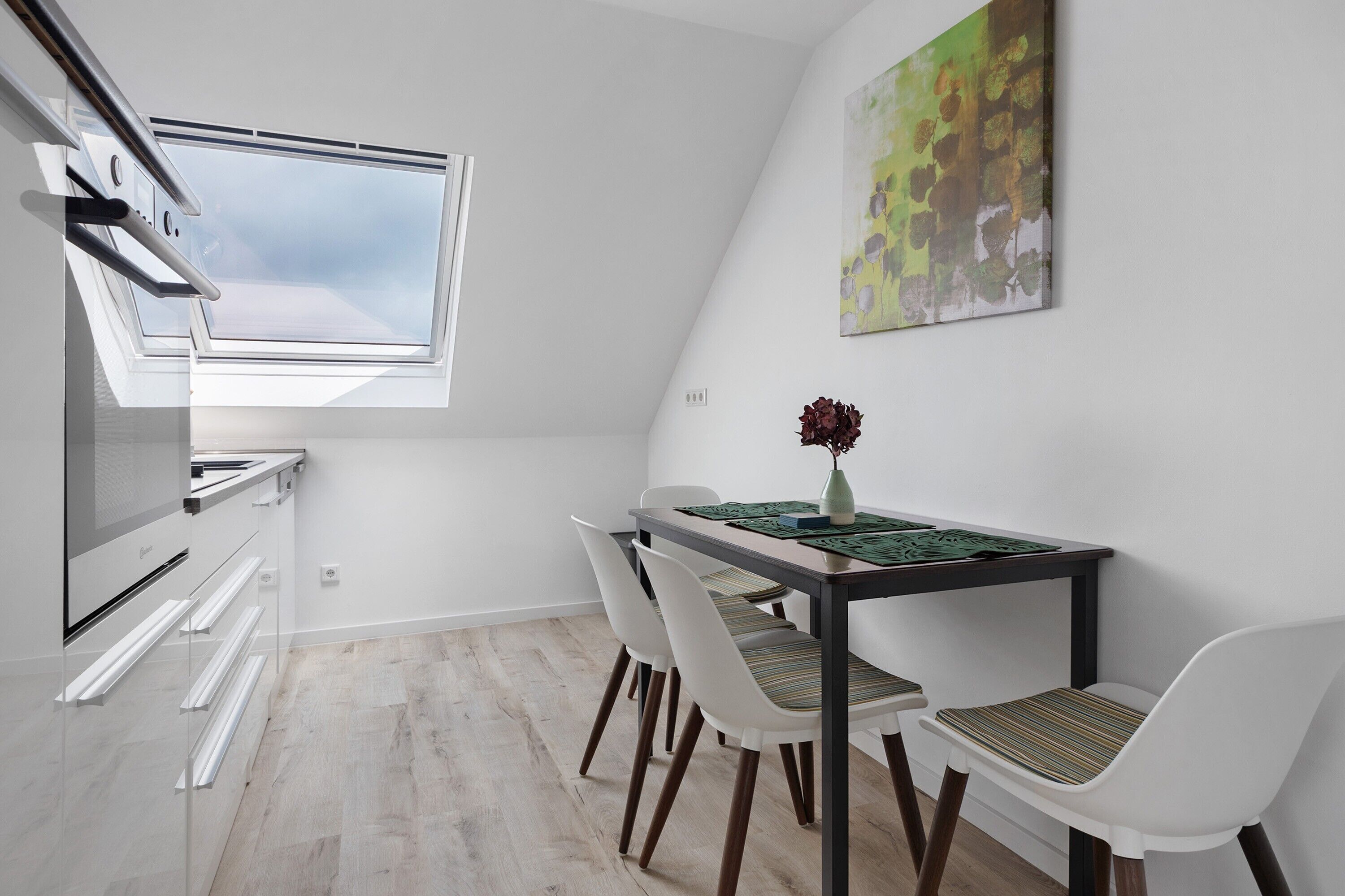 A compact kitchen with a skylight, a dining table with chairs, and a single vase with flowers.