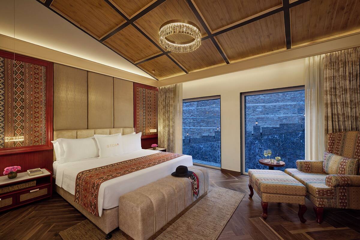 A spacious bedroom with a large bed, a sitting area with a chair, and a view of the city through large windows.