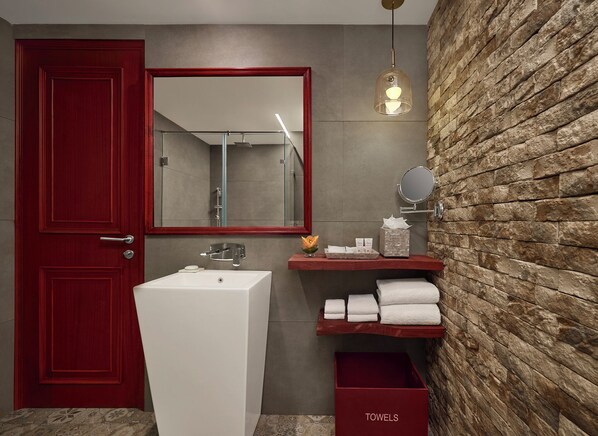 A bathroom with a red door, a white sink, a mirror, a towel rack, and a stone wall.