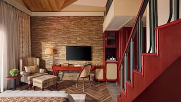A hotel room with a stone wall, a TV, a desk, a chair, a bed, and a staircase.