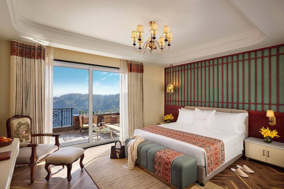 A spacious bedroom with a large bed, a chandelier, a view of the mountains, and a balcony with outdoor furniture.