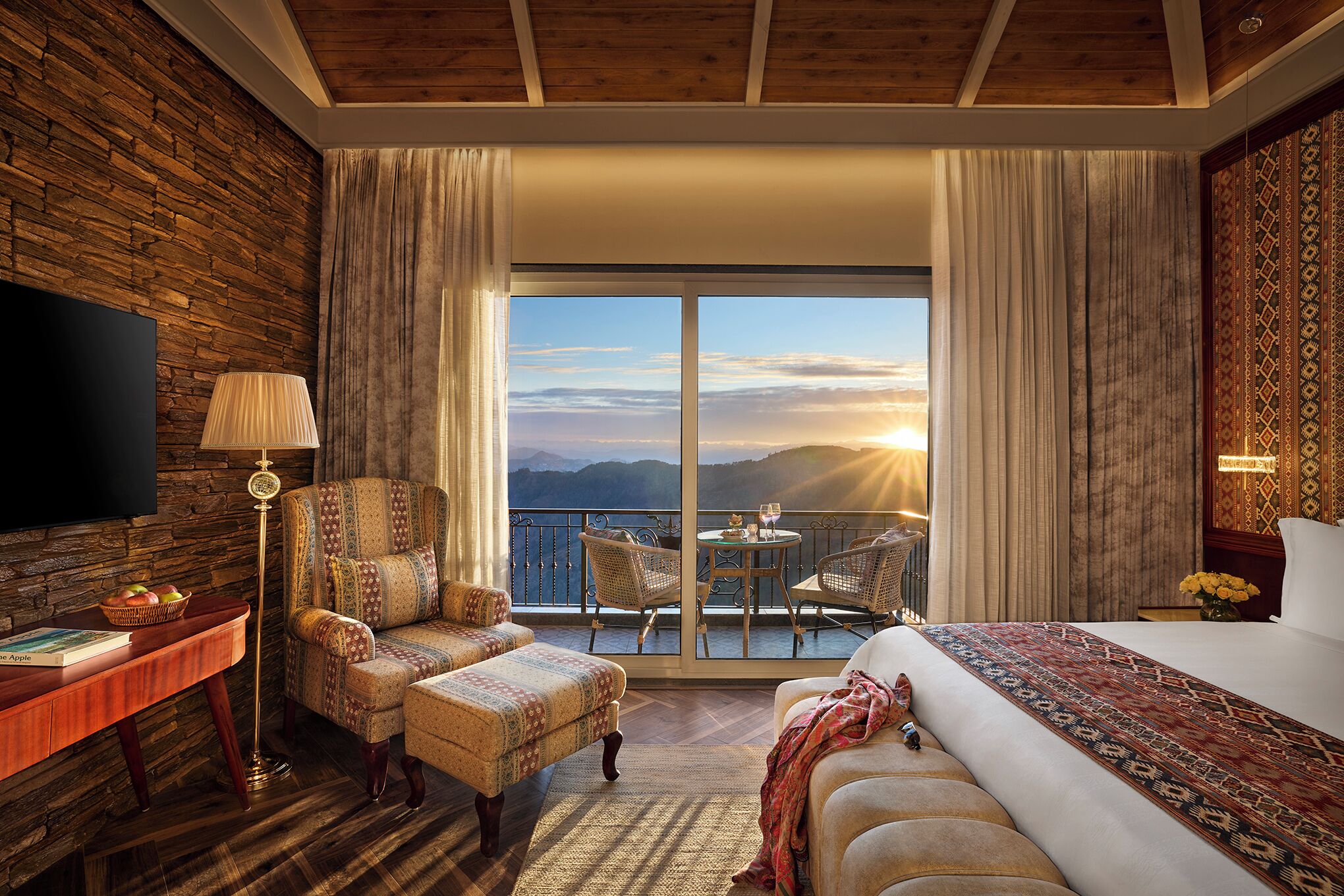 Habitación de hotel con una cama grande, televisión, escritorio con lámpara, silla, pequeña mesa con sillas y balcón con vistas a las montañas.