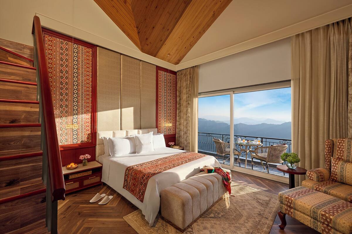 A spacious bedroom with a large bed, a balcony with a view of the mountains, and a wooden ceiling.