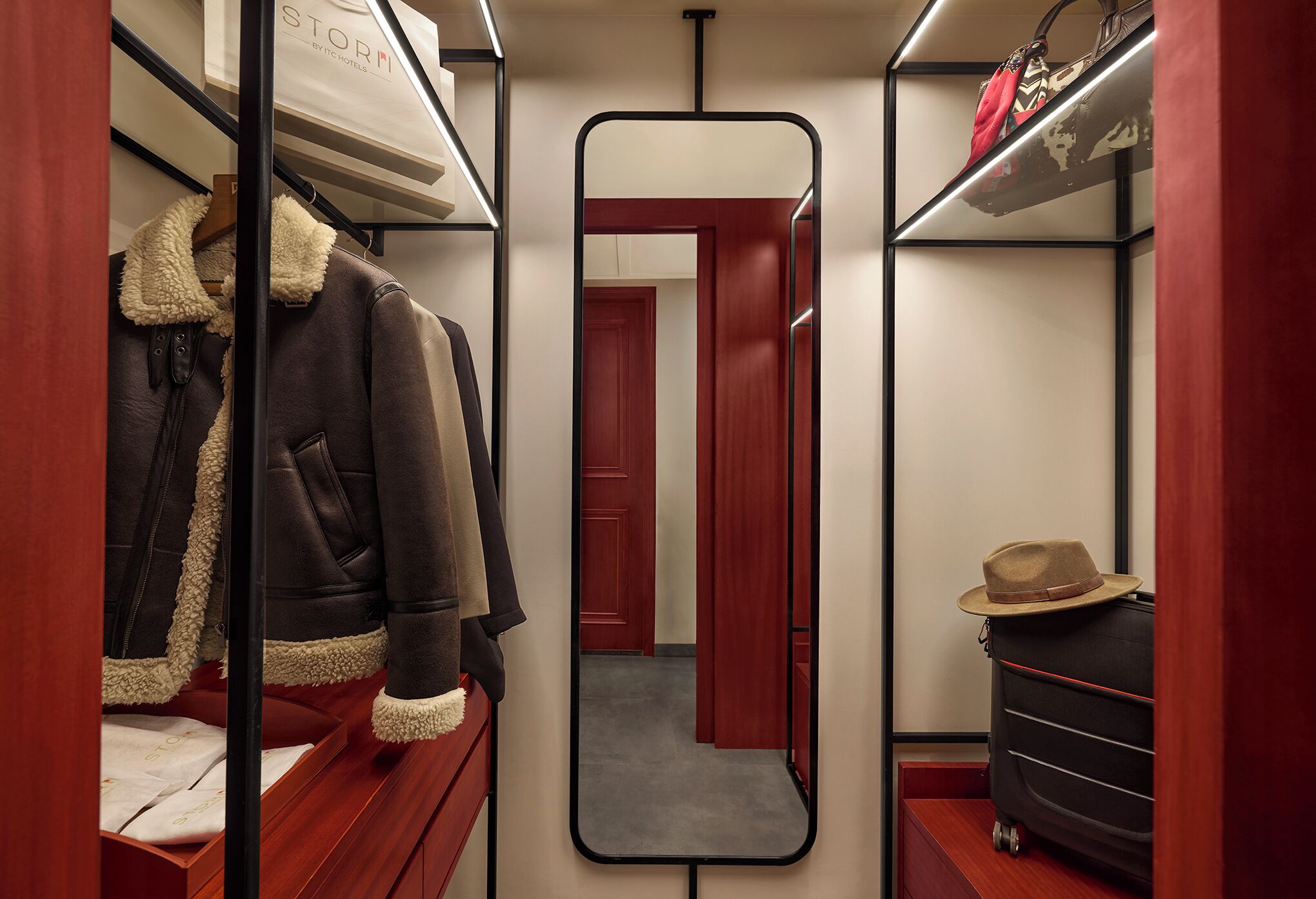 A narrow walk-in closet with mirrored doors, wooden shelving, and a red wall.