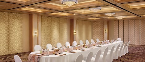 A banquet hall with a long table set for a formal event, featuring white chairs and a patterned carpet.