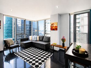 Een moderne woonkamer met een zwart-wit geruit tapijt, een grijze hoekbank, een kleine tafel met een bloevas, en grote ramen met uitzicht op de stad.