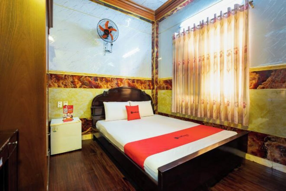 Une chambre d’hôtel avec un lit en bois, une petite table de chevet, un ventilateur fixé au mur et une fenêtre avec des rideaux.