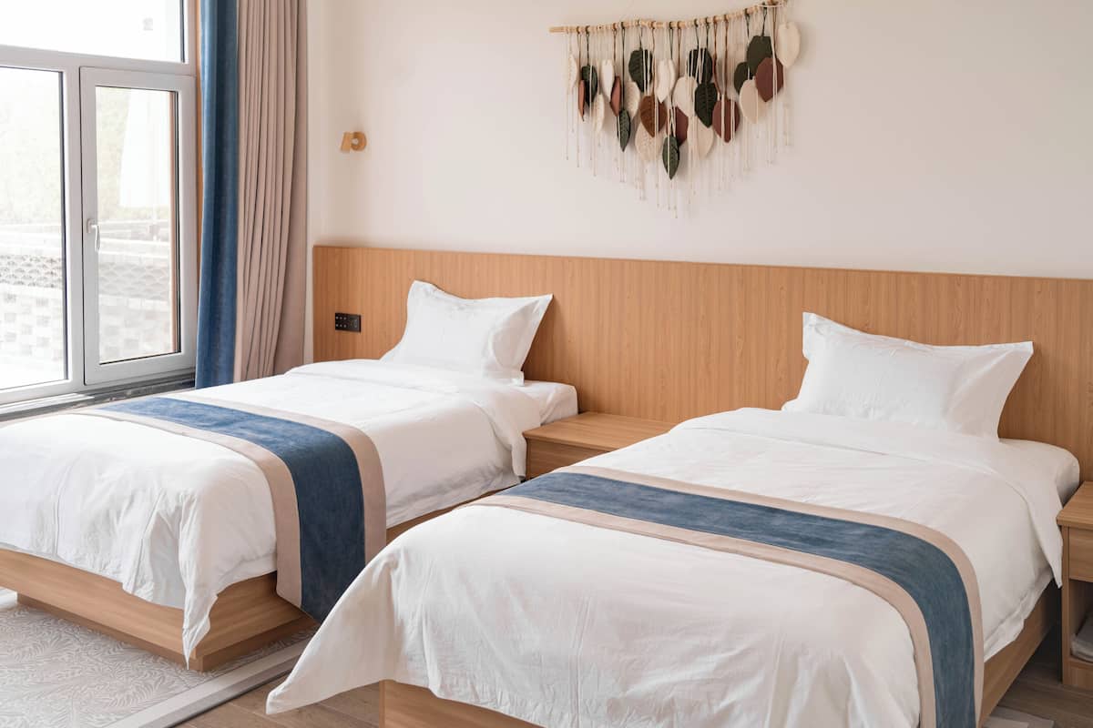 Habitación de hotel con dos camas, un cabecero de madera, un adorno de macramé en la pared y un ventanal con cortinas.