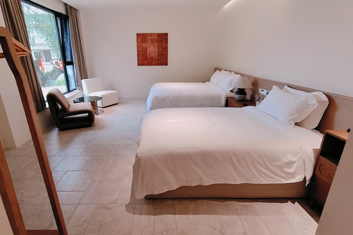Habitación de hotel con dos camas, una silla, una mesita y un cuadro en la pared.