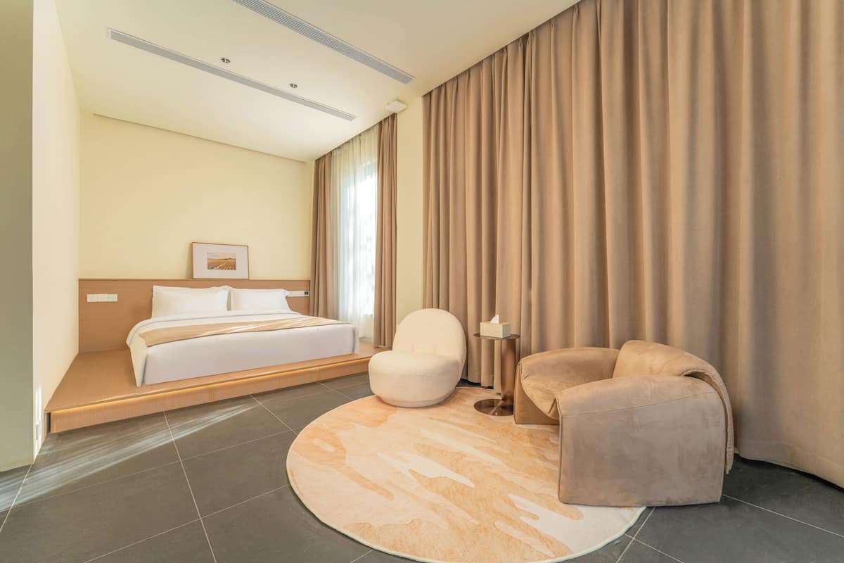 Una habitación de hotel moderna con una cama, un tapete redondo color beige, un sillón blanco y un sofá.