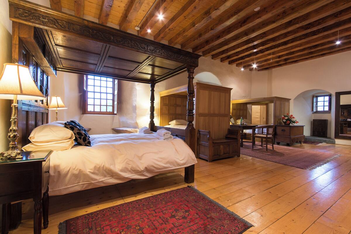 A traditional bedroom with a four-poster bed, wooden ceiling beams, and a large mirror.