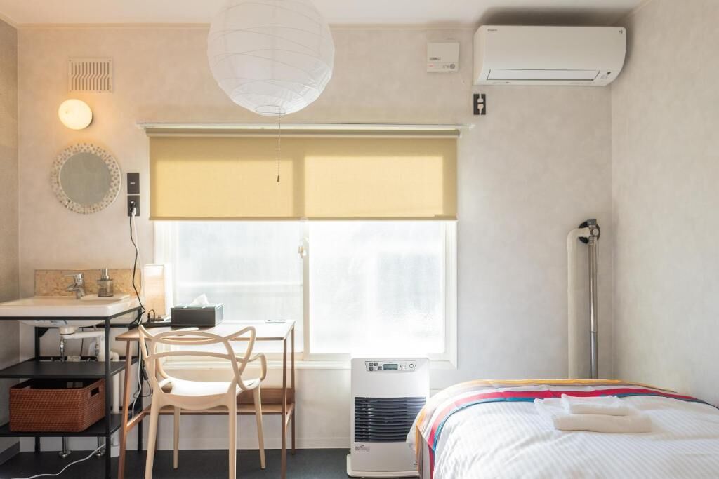 A hotel room with a bed, a desk with a chair, a window with blinds, a bedside table, a lamp, and an air conditioner.