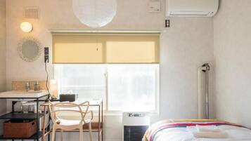 A hotel room with a bed, a desk with a chair, a window with blinds, a bedside table, a lamp, and an air conditioner.
