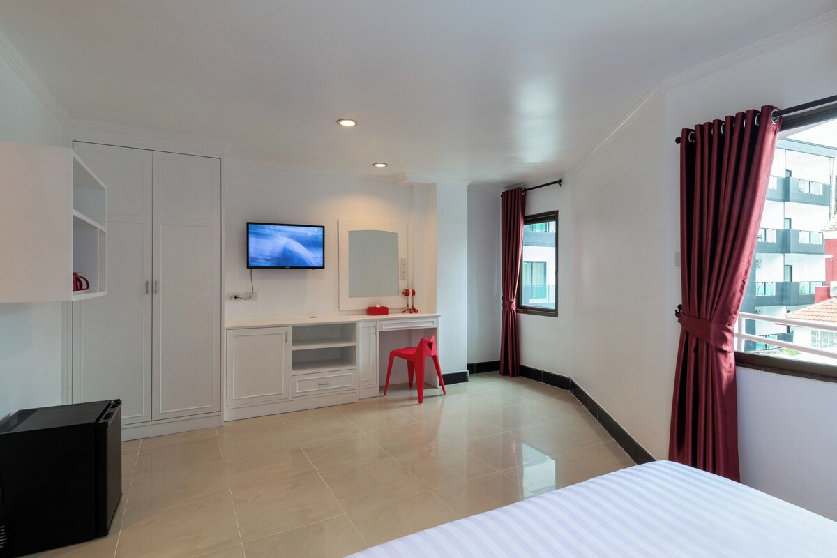 Una habitación de hotel con cama, escritorio, televisor, una silla roja y ventana con cortinas.