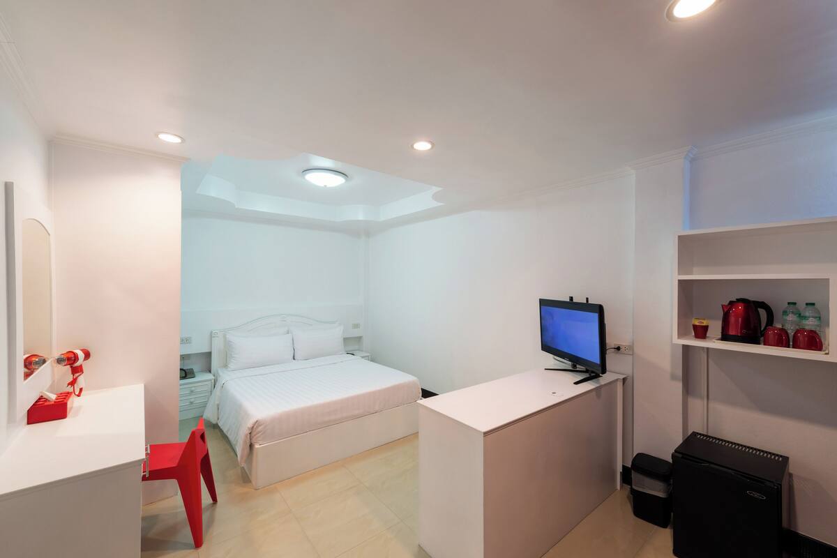 Ein kleines Hotelzimmer mit Bett, Schreibtisch mit Computer, rotem Sessel, einer kleinen Küchenzeile und Fernseher.