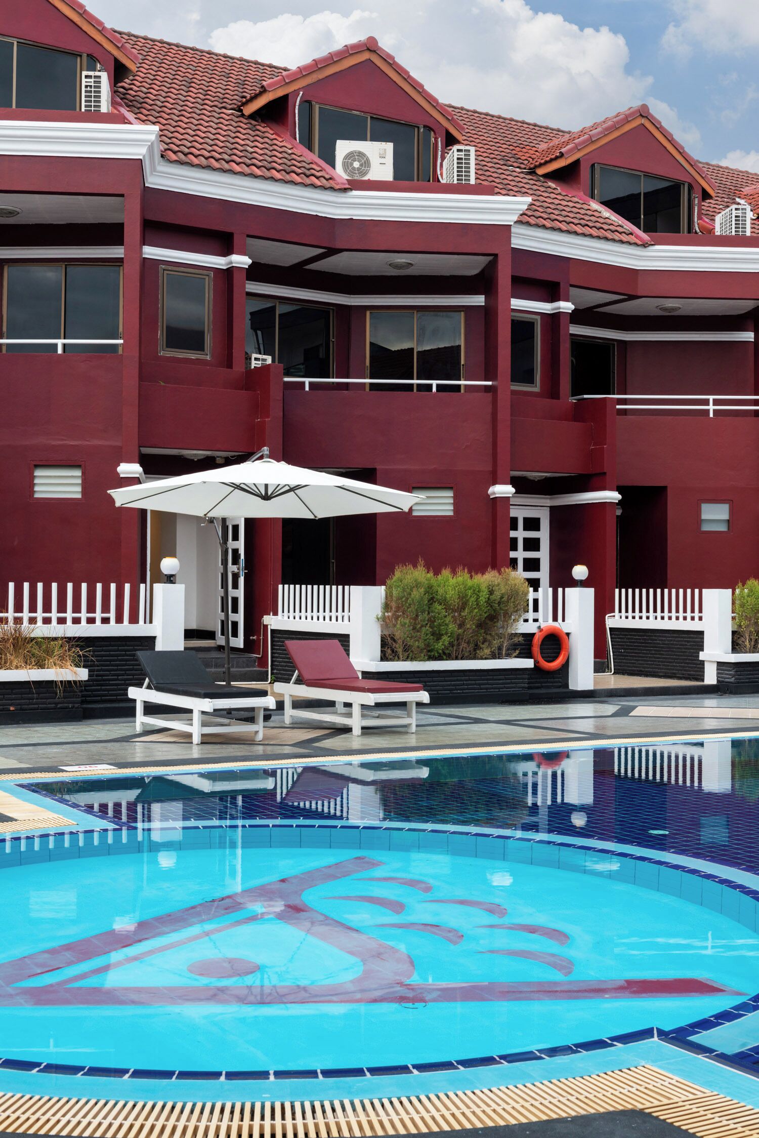 Une piscine avec des chaises longues et des parasols, un bâtiment rouge doté de nombreuses fenêtres et de balcons, et une clôture blanche.