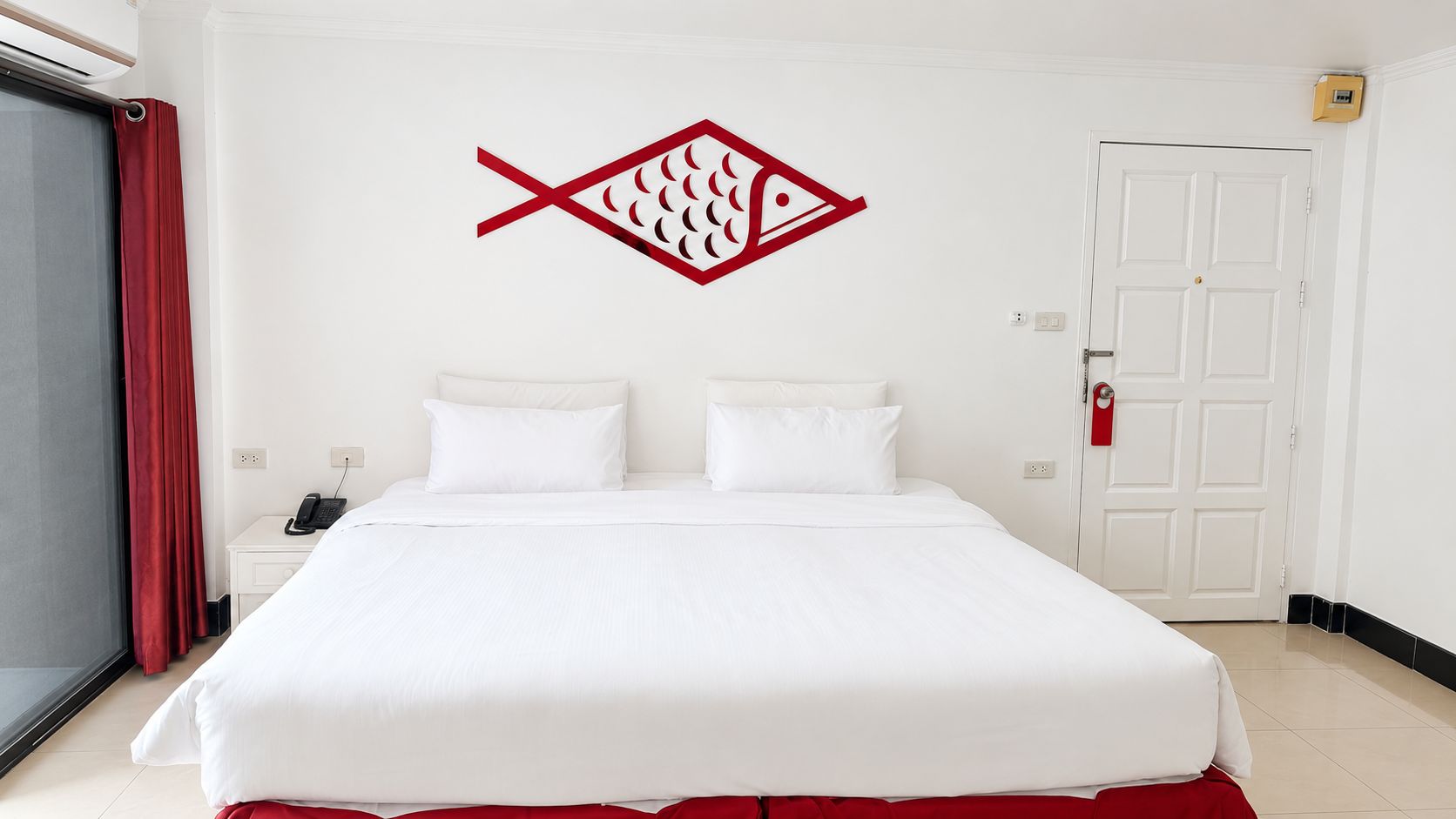 Habitación de hotel con una cama grande, una cortina roja, un símbolo de pez montado en la pared, una puerta blanca y un teléfono en la mesita de noche.
