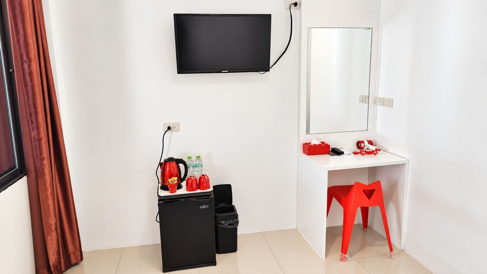 A hotel room with a flat-screen TV mounted on the wall, a desk with a mirror, a red chair, and a small black storage unit with red kettles and bottles.