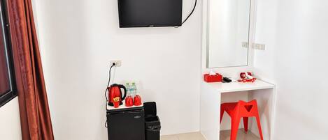 A hotel room with a flat-screen TV mounted on the wall, a desk with a mirror, a red chair, and a small black storage unit with red kettles and bottles.