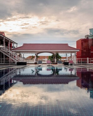 Une piscine avec une terrasse carrelée, un espace de repos couvert et, au fond, un bâtiment avec un toit rouge.