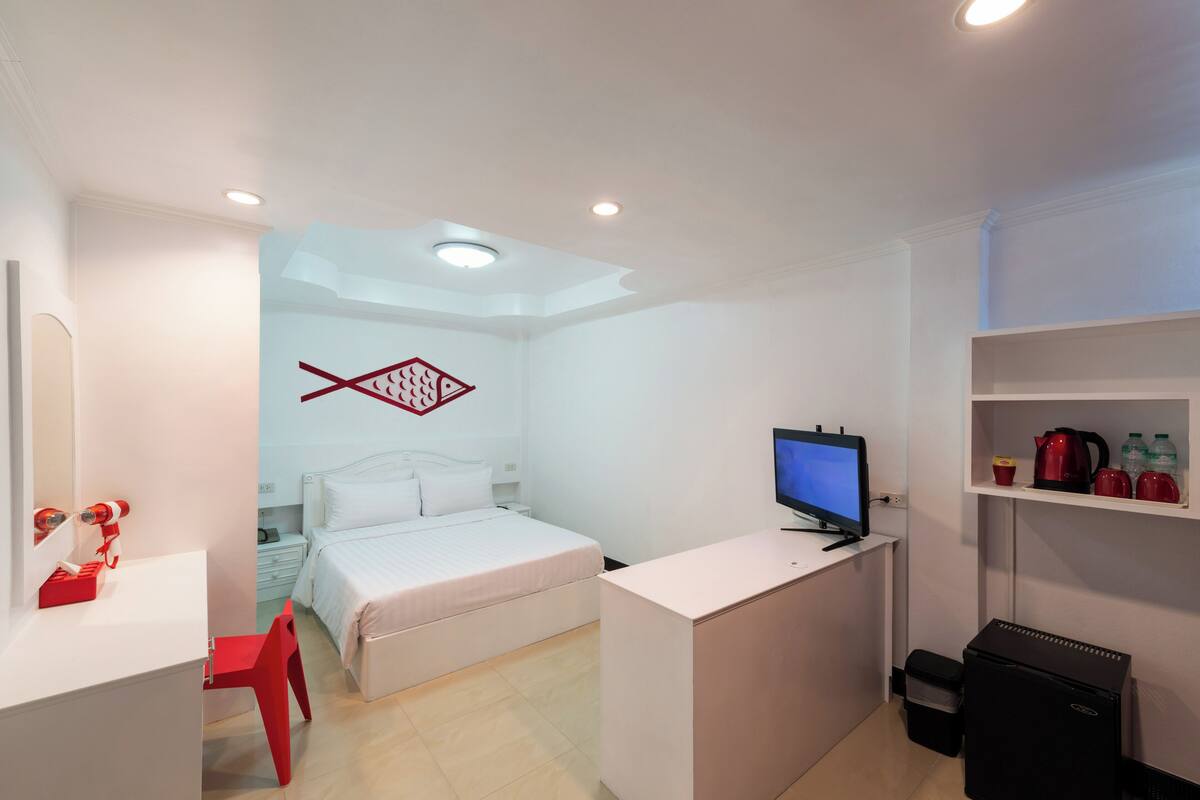 Ein kleines Hotelzimmer mit Bett, Schreibtisch mit Computer, rotem Sessel, einer kleinen Küchenzeile und einem Fernseher.