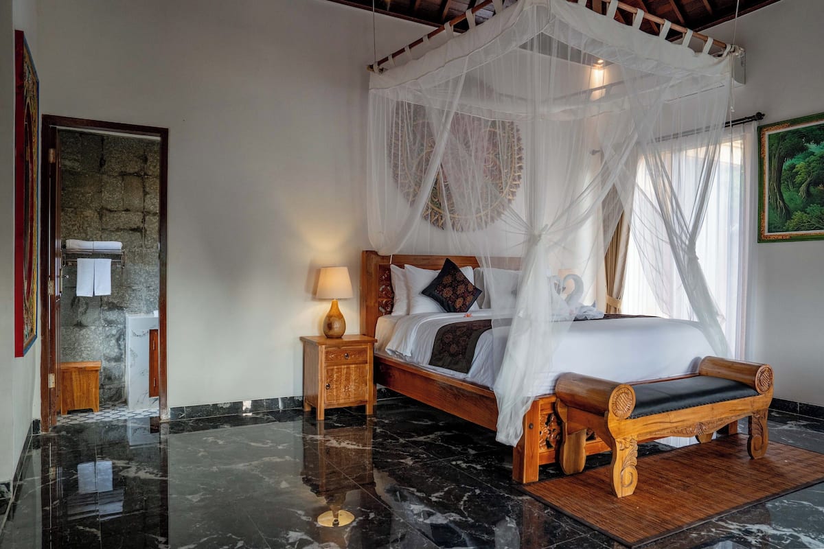A bedroom with a bed under a canopy, a wooden bench, a nightstand with a lamp, and a stone wall.