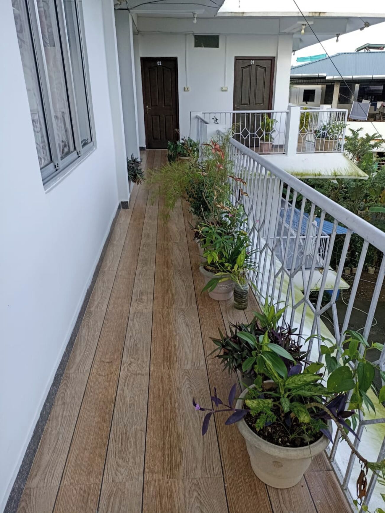 A wooden deck with potted plants, a white railing, and a building with two doors in the background.