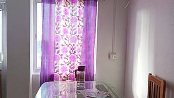 A dining area with a glass table, two chairs, a window with purple curtains, and an air conditioning unit above.