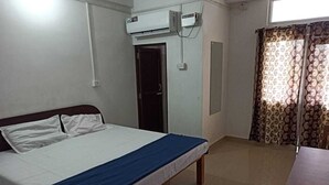 A bedroom with a bed, an air conditioning unit, a door, a window with curtains, and a small table.