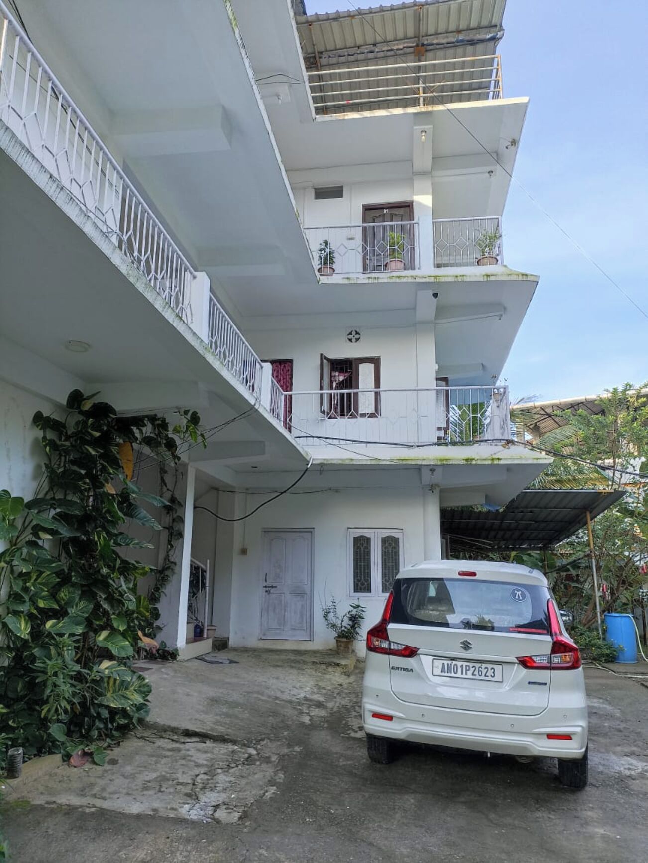 A multi-story residential building with a white exterior, a parked car, and a balcony with potted plants.