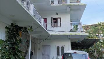 A multi-story residential building with a white exterior, a parked car, and a balcony with potted plants.