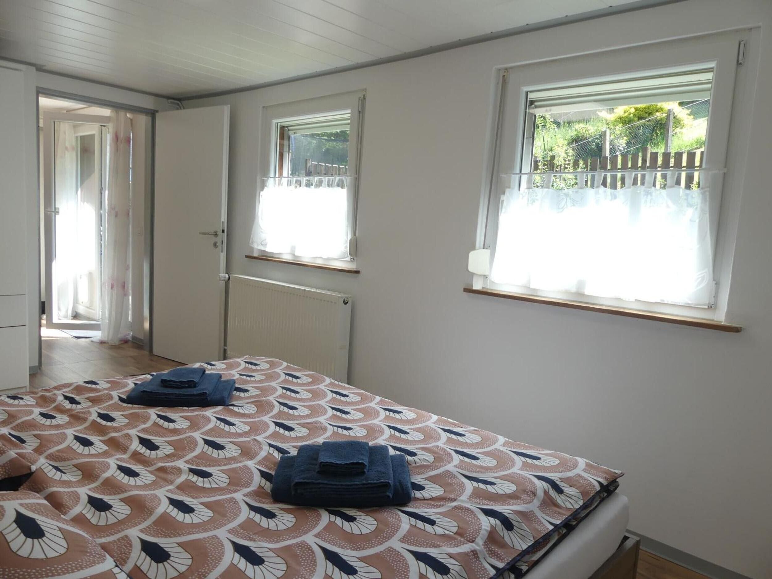 A room with a bed, two windows, a radiator, and a door leading to another room.