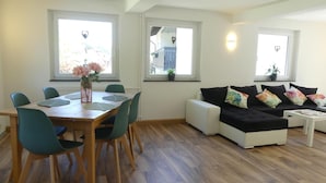 Ein modernes Wohnzimmer mit Essbereich, großen Fenstern und einer Couch mit bunten Kissen.
