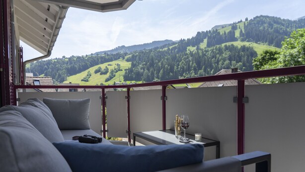 A balcony with a sofa, a small table with a wine glass, and a view of green hills.