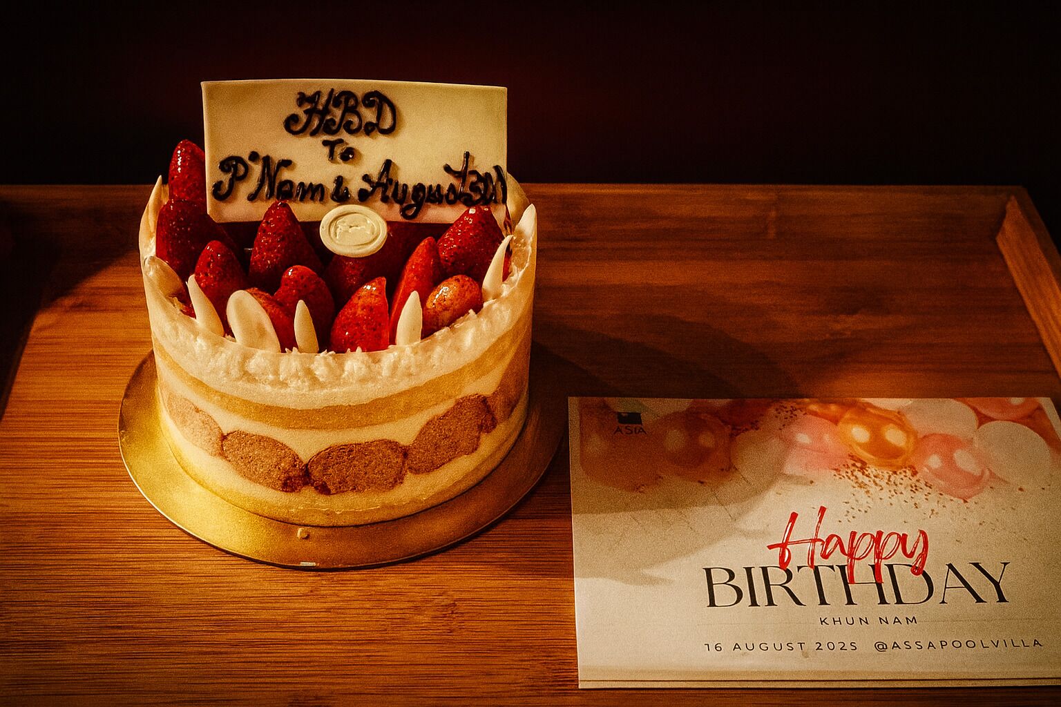A birthday cake with a handwritten card and a birthday card on a wooden table.