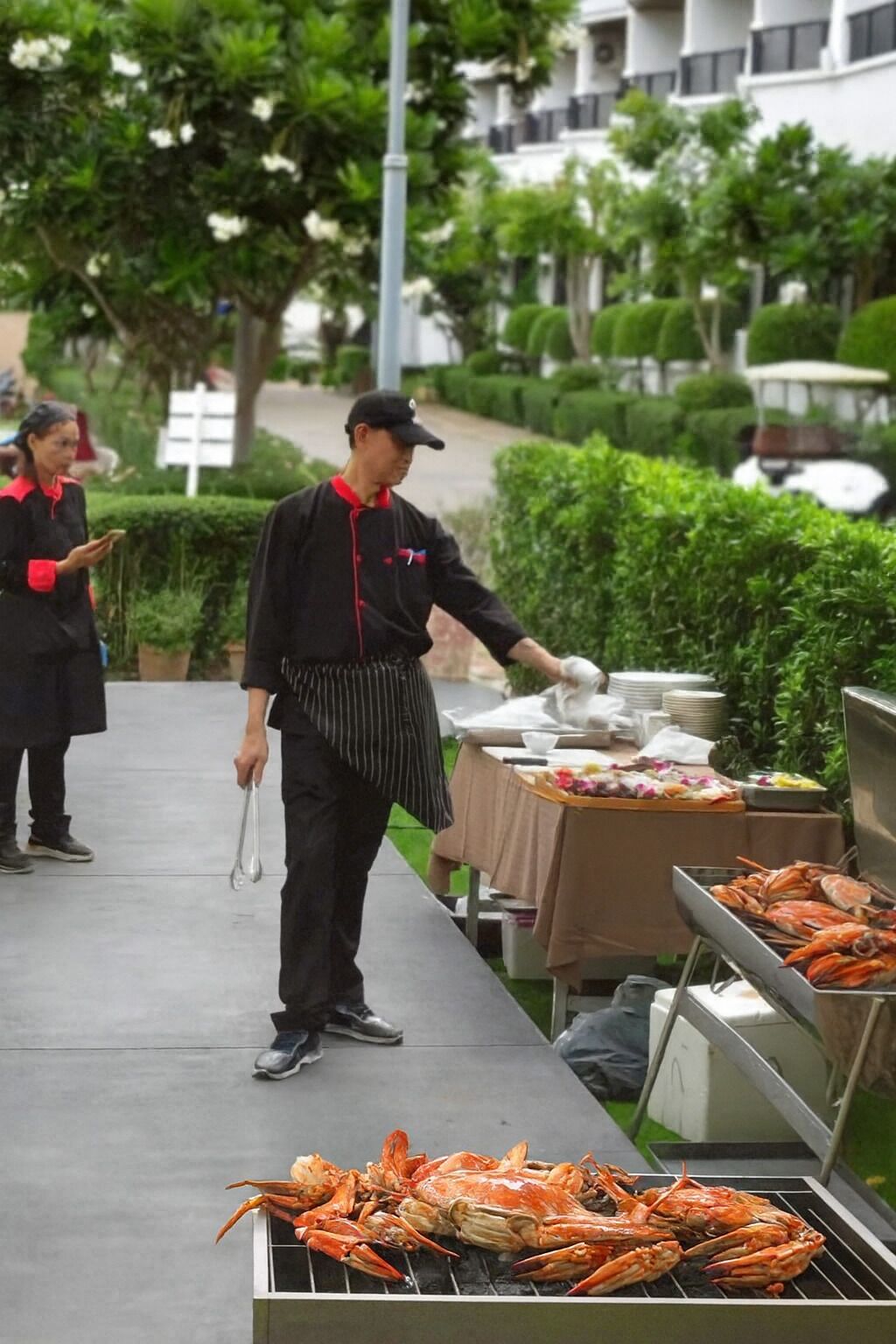 Iemand barbecuet krabben buiten, met een tafel vol gerechten en een bord in de achtergrond.