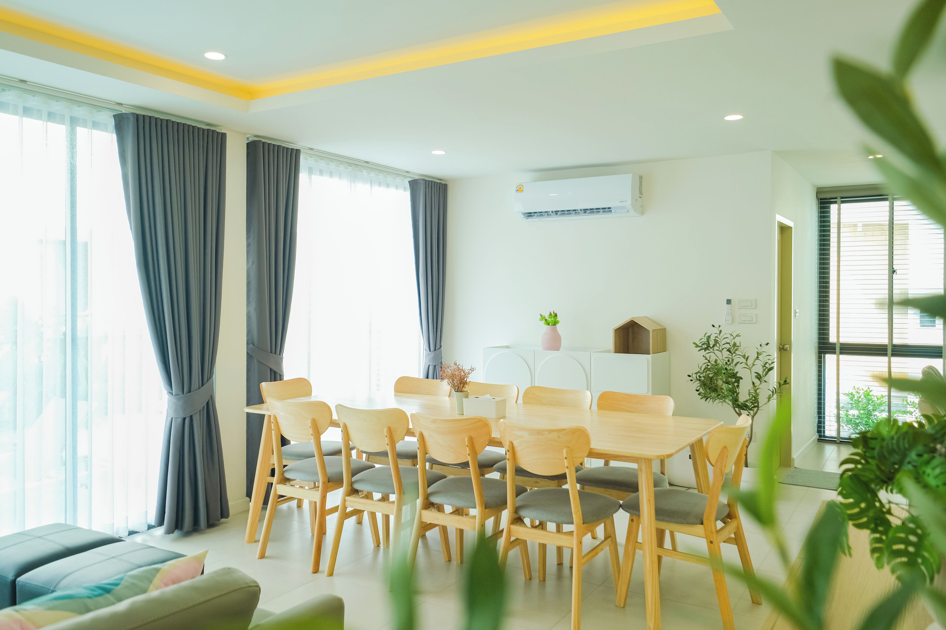 A bright, modern dining area with a wooden table and chairs, large windows with curtains, and a white air conditioning unit on the wall.