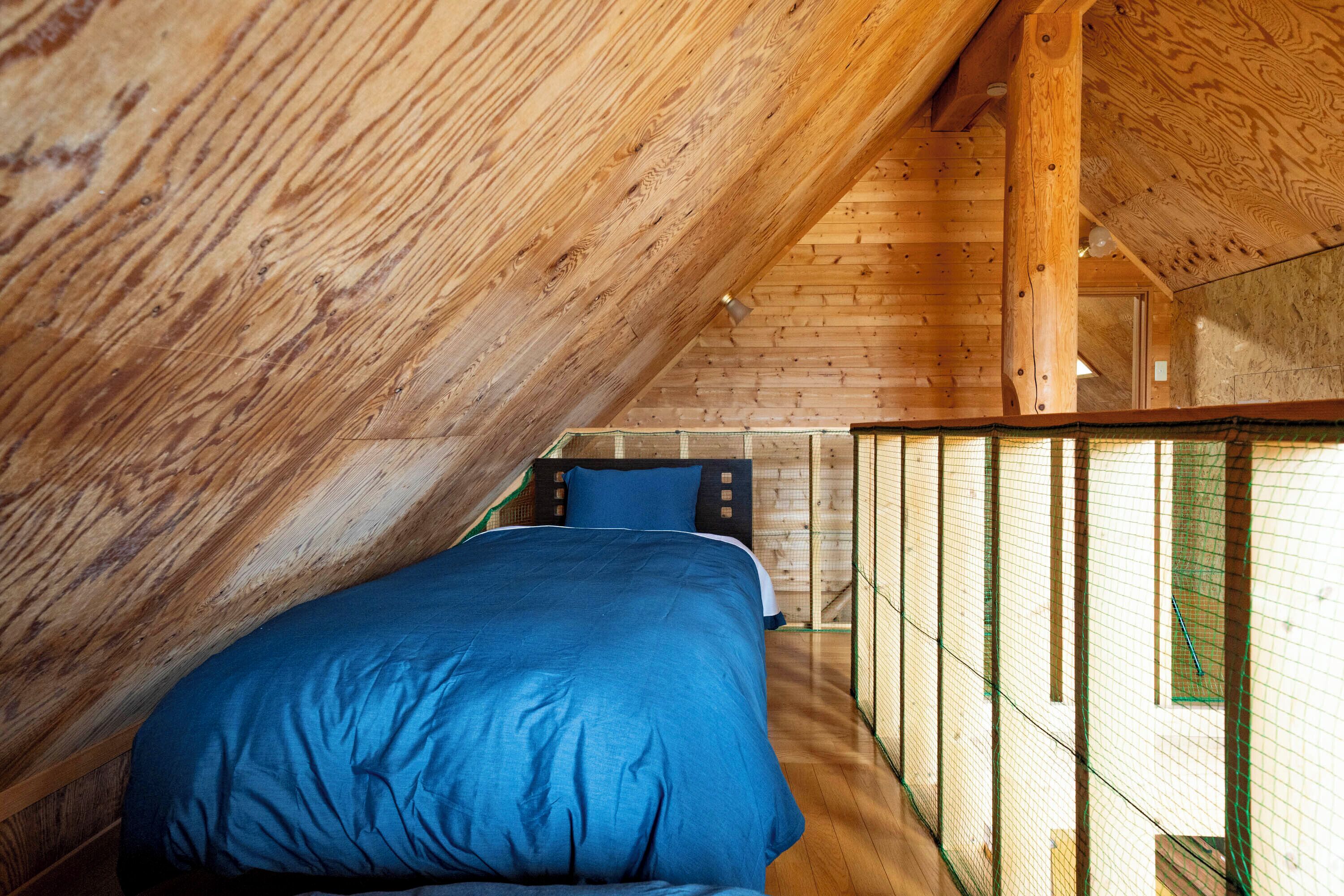Un dormitorio con techo inclinado, paredes de madera, cama con ropa de cama azul y vista a través de una ventana con mosquitero verde.