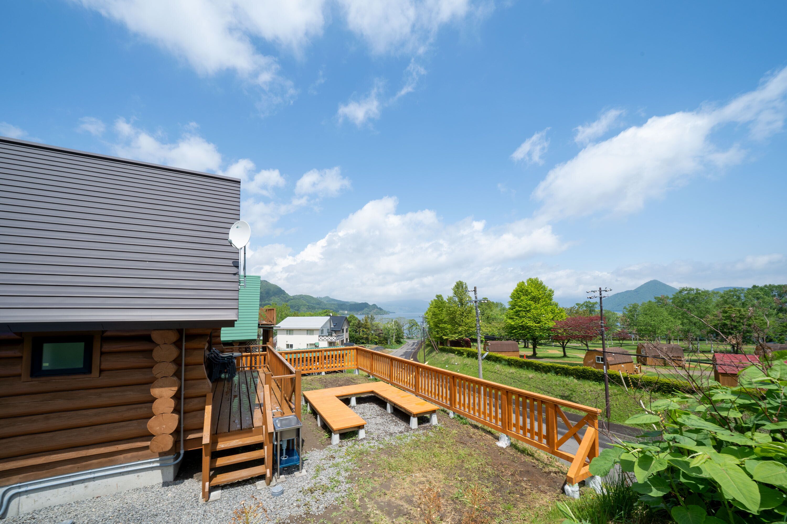 A wooden cabin with a deck and outdoor seating area, surrounded by greenery and mountains in the background.