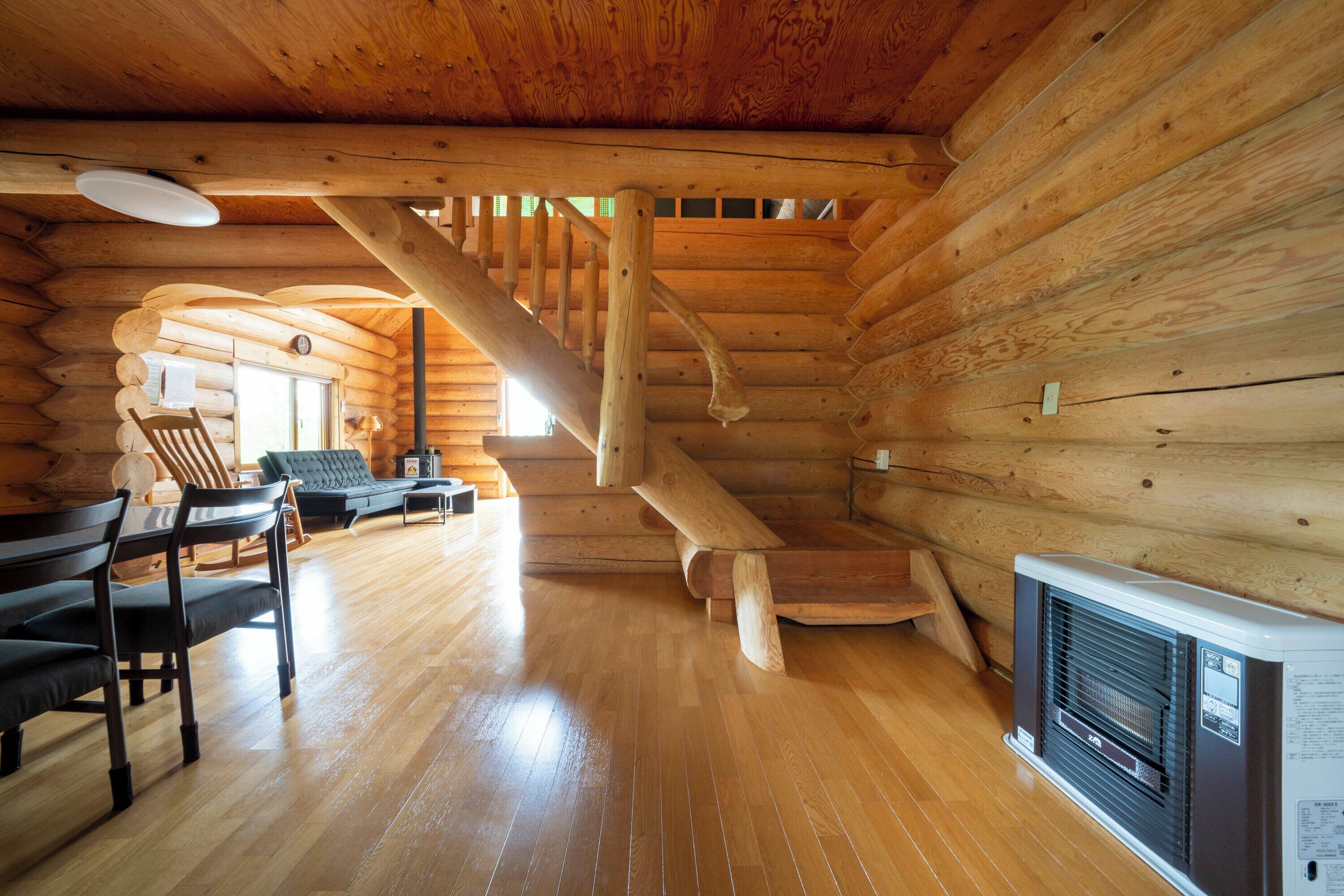A log cabin interior with a wooden staircase, a fireplace, and a dining area with chairs and a table.