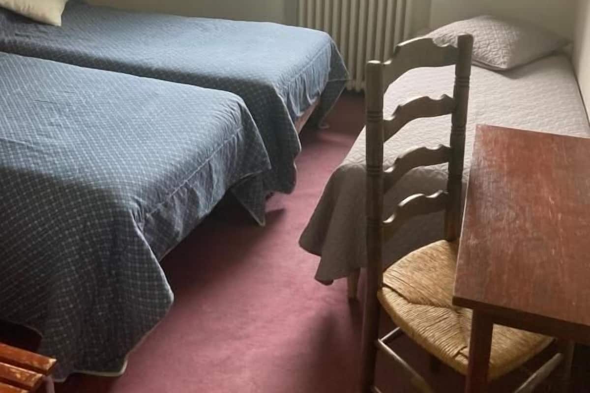 A bedroom with two beds, a wooden chair, a table, and a window with a view of a car and greenery outside.