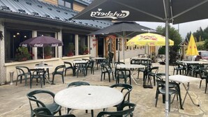 An outdoor patio area with round tables and chairs, umbrellas, and a building with a metal roof. - HOTEL LA GRANGE (Boutx)