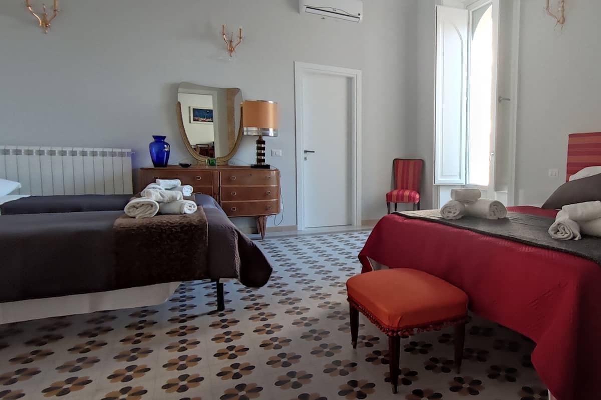 A hotel room with two beds, a red and black striped headboard, a red chair, a small table, a mirror, a lamp, and a patterned floor.