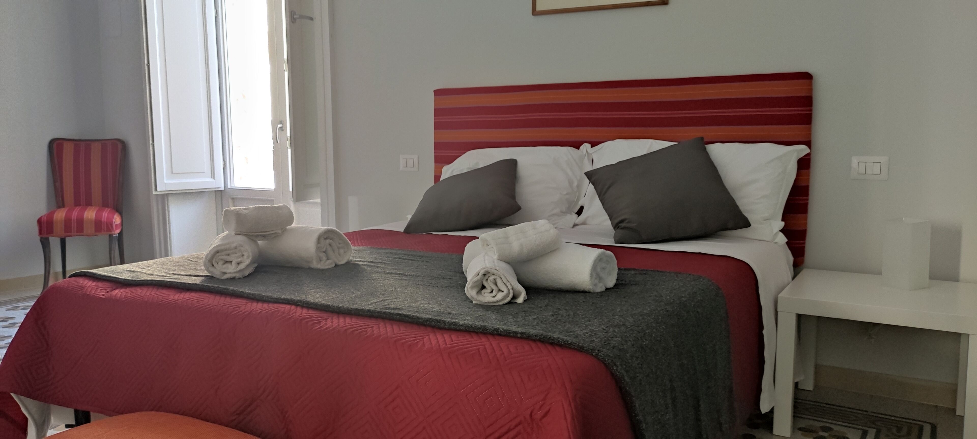 A neatly made bed with pillows and rolled towels, a red upholstered chair, and a white nightstand.