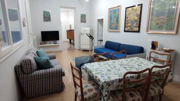 Un espace de vie compact comprenant un canapé, une table à manger avec des chaises, un téléviseur, un réfrigérateur et des œuvres d’art accrochées aux murs.