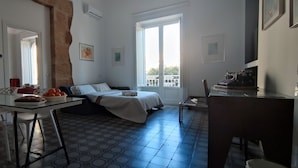 A compact room with a bed, a small table with fruits, a chair, and a window with a balcony view. - Palazzo Caratozzolo (Agrigento)