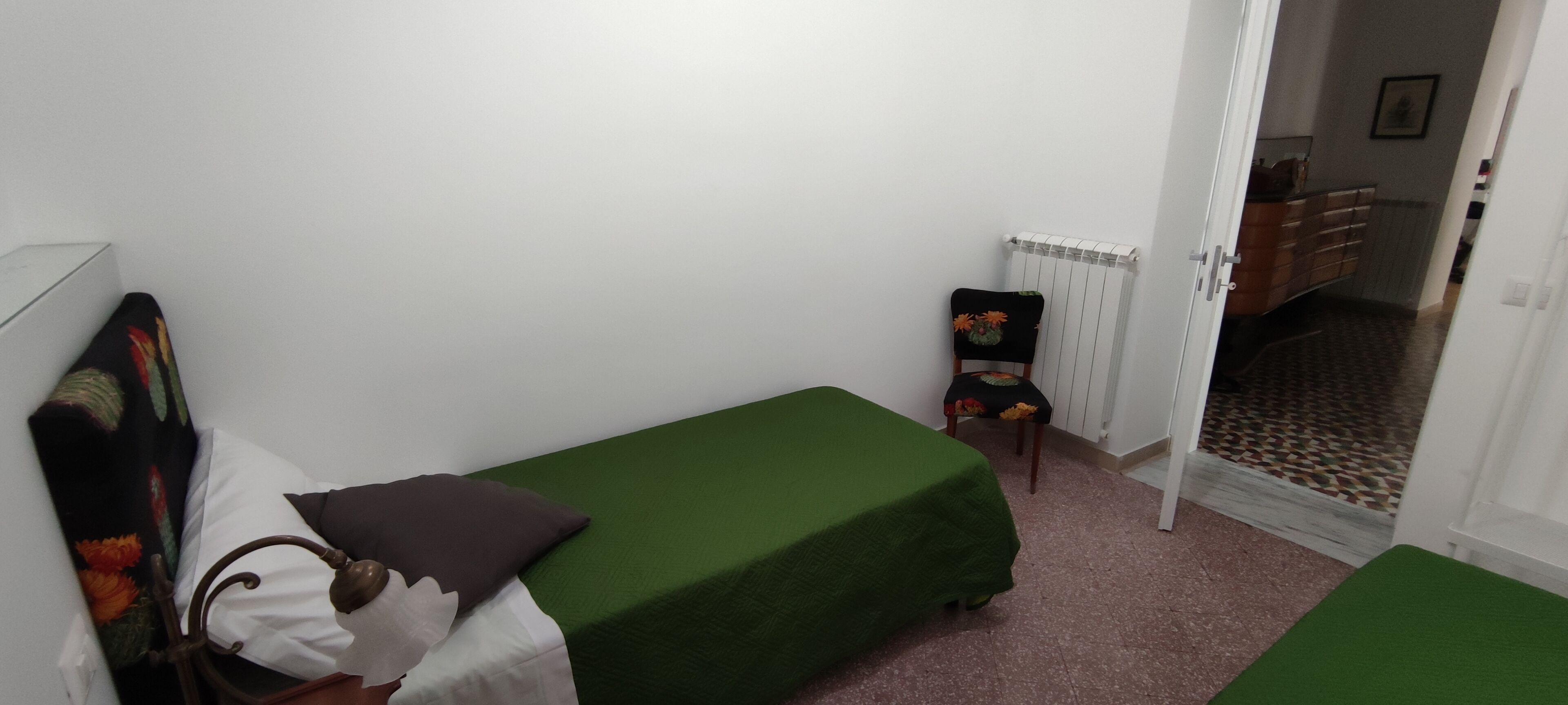 A small, single-bed room with a green bedspread, a chair with a floral cushion, and a radiator.