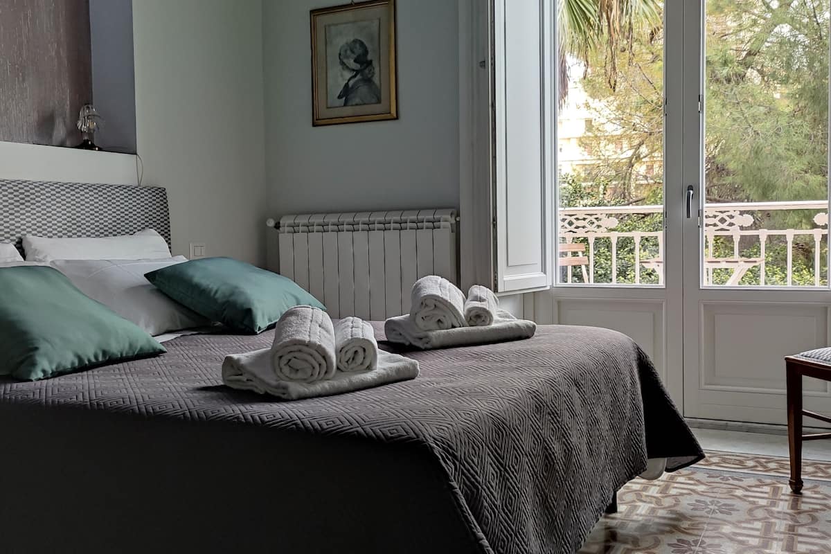 A neatly made bed with a patterned bedspread, pillows, and rolled towels. A chair is positioned near a window with a view of greenery outside.