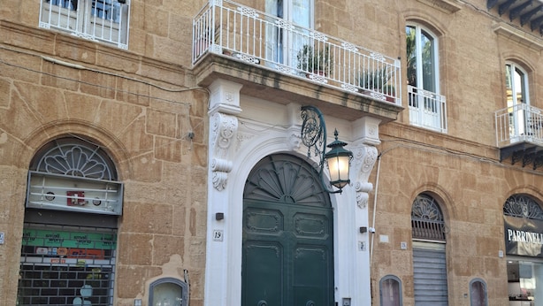 A building with a green door, a balcony with white railings, and a small shop on the ground floor. - Palazzo Caratozzolo (Agrigento)