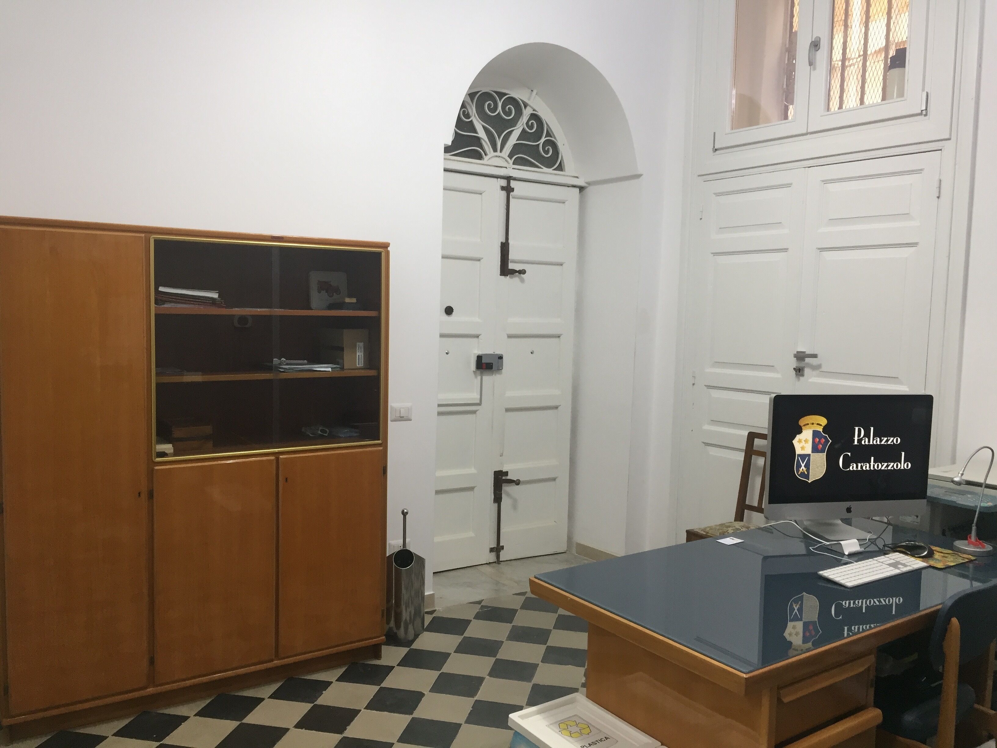 A room with a checkered floor, a wooden cabinet, a desk with a computer, and a door with a decorative arch.